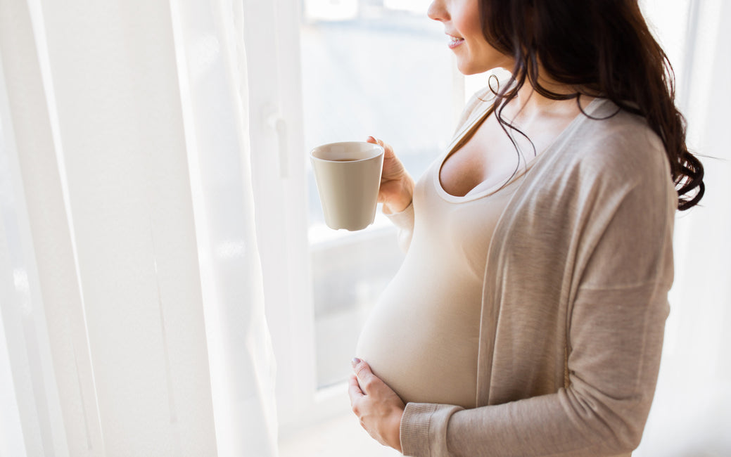 Pregnant woman standing at the window and drinking coffee