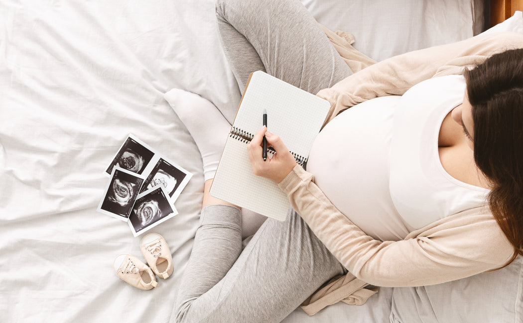 Pregnant woman looks at ultrasound images and writes baby names in a notebook.