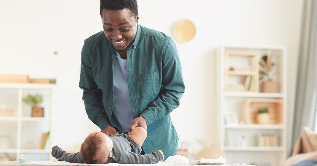 A father stands in front of the changing table, entertaining his baby while getting dressed.