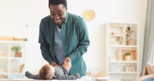 A father stands in front of the changing table, entertaining his baby while getting dressed.