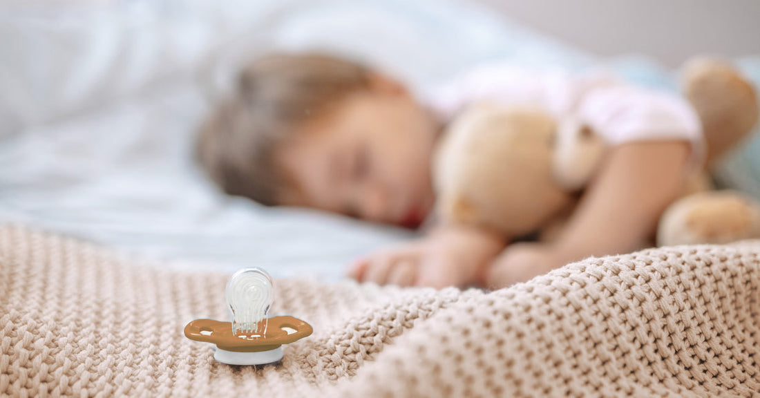 Baby is lying in bed and sleeping. In the foreground, there is a pacifier.