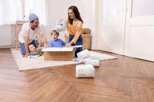 Mother, father, and two small children are unpacking a LILLYDOO diaper box together. One child is sitting inside the box. Diaper packs are on the floor.