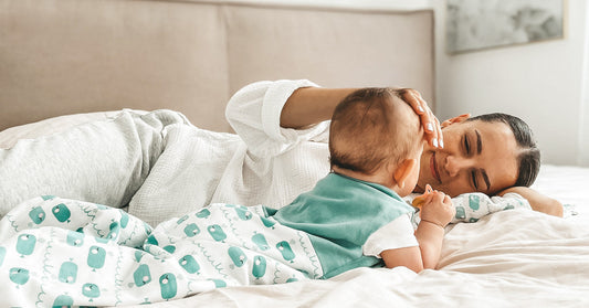 A young woman is lying on a bed with a baby in a sleeping bag, gently stroking its head.