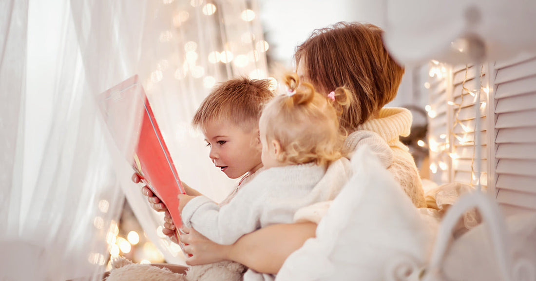 A mother sits with her two children on her lap in a cozy, softly lit room. They are holding a red notebook in their hands.