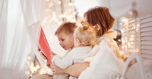 A mother sits with her two children on her lap in a cozy, softly lit room. They are holding a red notebook in their hands.